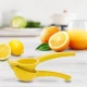Χρήση του κίτρινου στύφτη λεμονιού χειρός Pedrini στον πάγκο της κουζίνας δίπλα σε φρέσκα λεμόνια, πορτοκάλια και ένα ποτήρι χυμό.