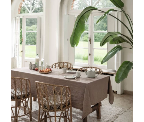 Υφασμάτινο τραπεζομάντηλο Finesse WOW σε γήινο καφέ χρώμα, στρωμένο σε ξύλινο τραπέζι με καρέκλες ρατάν σε φωτεινό χώρο με φυτά.