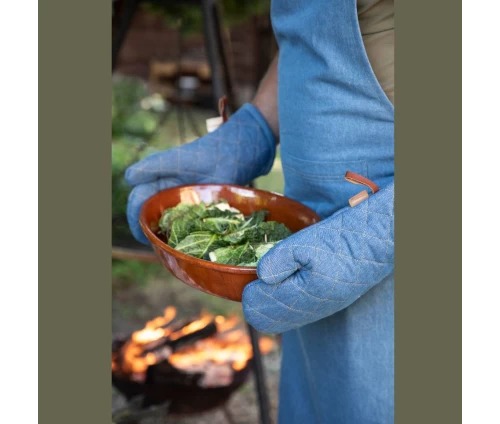 Άντρας φοράει τζιν ποδιά και δύο τζιν γάντια φούρνου Finesse CHEF, κρατώντας ένα πήλινο σκεύος με φαγητό μπροστά από αναμμένη φωτιά (BBQ).