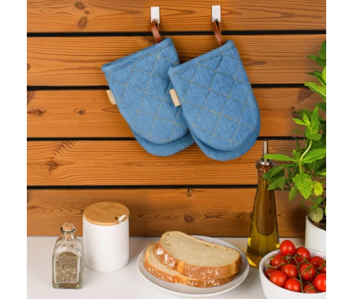 Δύο τζιν μίνι γάντια κουζίνας Finesse CHEF κρεμασμένα πρακτικά σε ξύλινο τοίχο, πάνω από πάγκο προετοιμασίας με φρέσκο ψωμί και ντοματίνια.