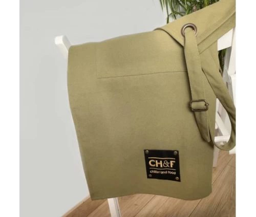 Λαδί ποδιά Finesse CHEF BBQ ριγμένη κομψά σε καρέκλα, εστιάζοντας στο μαύρο δερμάτινο patch με το λογότυπο 